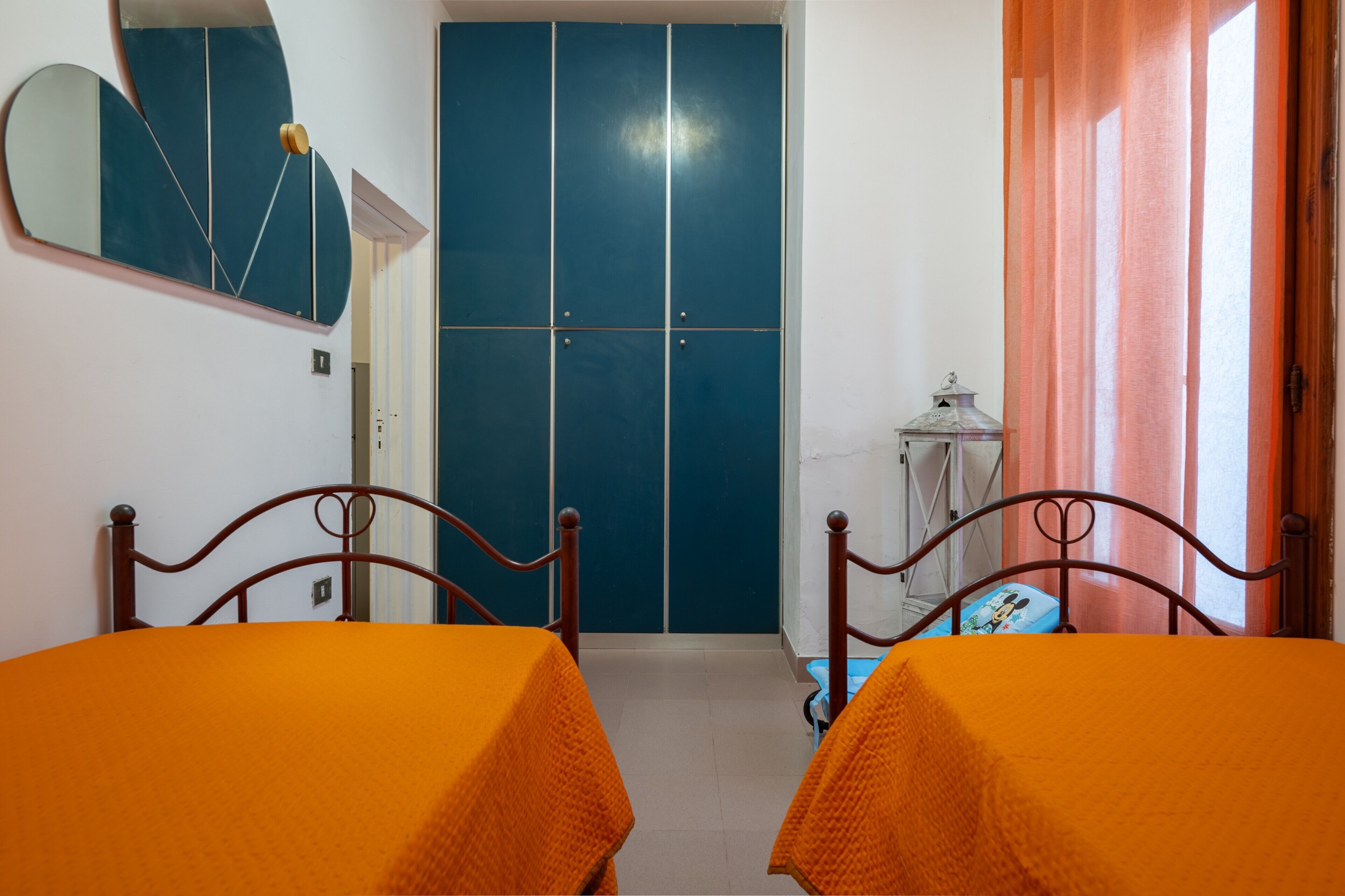 A room with two beds, a wardrobe, a mirror, and a small table with a lamp.