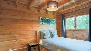 Un dormitorio de cabaña de madera con una cama, una mesita de noche, una lámpara, una ventana y un cuadro en la pared.