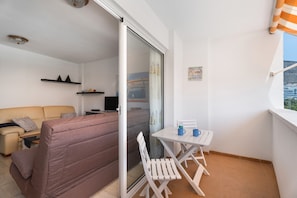 En balkong med ett bord och stolar, en soffa, en tv och en skjutdörr i glas som leder ut till en utsikt över staden.