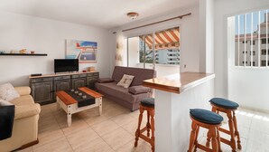A modern living room with a sofa, coffee table, TV, and a balcony with a striped awning. - Holiday Apartment 'Central Los Cristianos' with Mountain View, Pool and Wi-Fi (Los Cristianos)