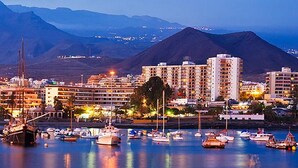 A marina with boats, a prominent building with multiple floors, and a mountainous backdrop. - Holiday Apartment 'Central Los Cristianos' with Mountain View, Pool and Wi-Fi (Los Cristianos)