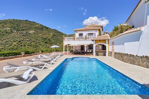 Une piscine avec des chaises longues, un espace patio et un bâtiment doté d’un balcon donnant sur un paysage vallonné.