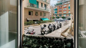 Vista dalla finestra su una strada con scooter parcheggiati e un edificio con balconi e un tendone verde.