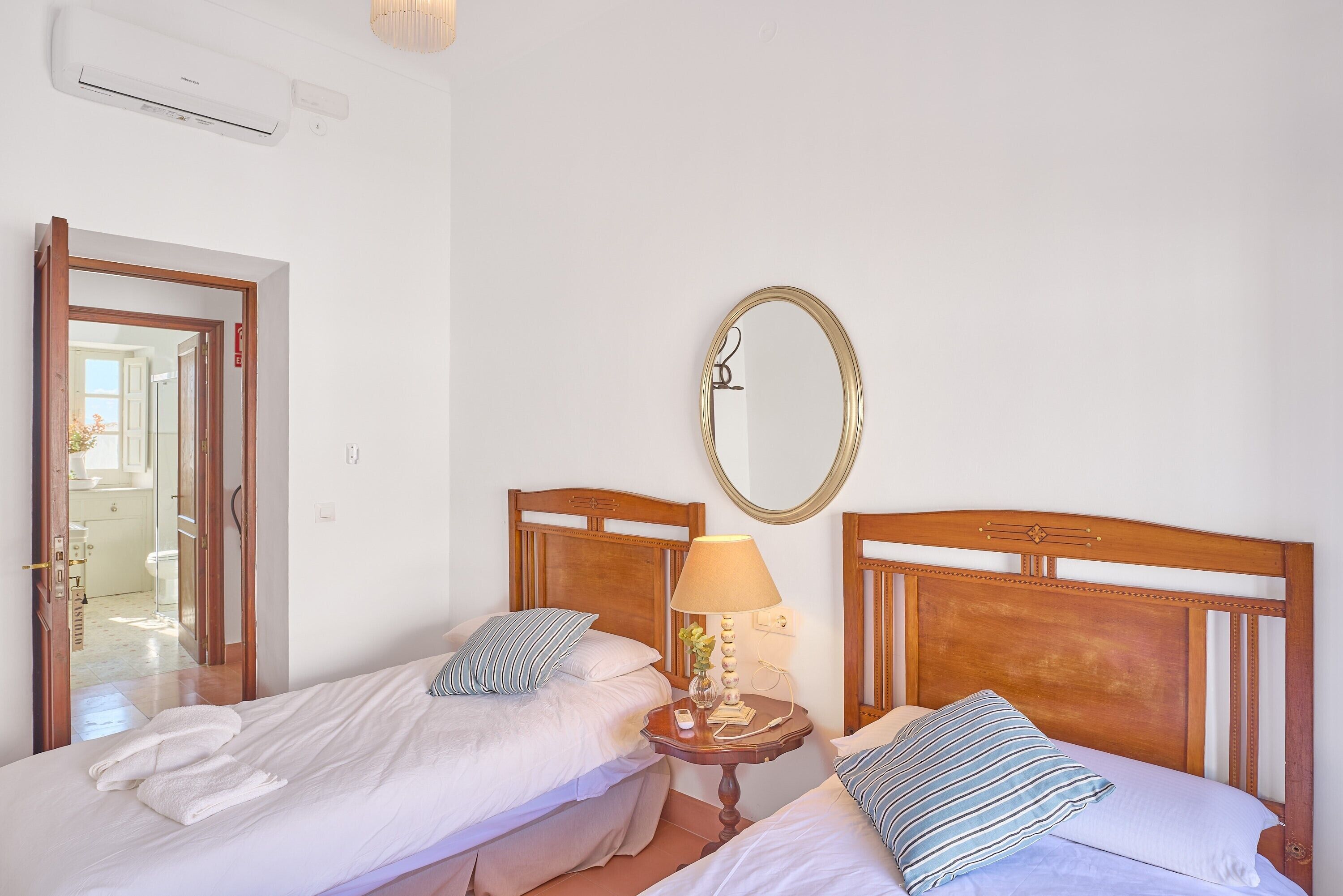 A hotel room with two beds, a bedside table with a lamp, a mirror, and an air conditioning unit.