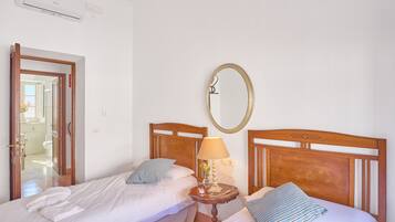 A hotel room with two beds, a bedside table with a lamp, a mirror, and an air conditioning unit.