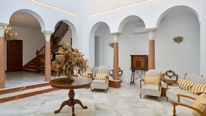 Eine geräumige Lobby mit Marmorböden, weißen Wänden und Bögen, ausgestattet mit einem runden Tisch, einer Blumenkomposition, mehreren Polsterstühlen und einer hölzernen Bank.