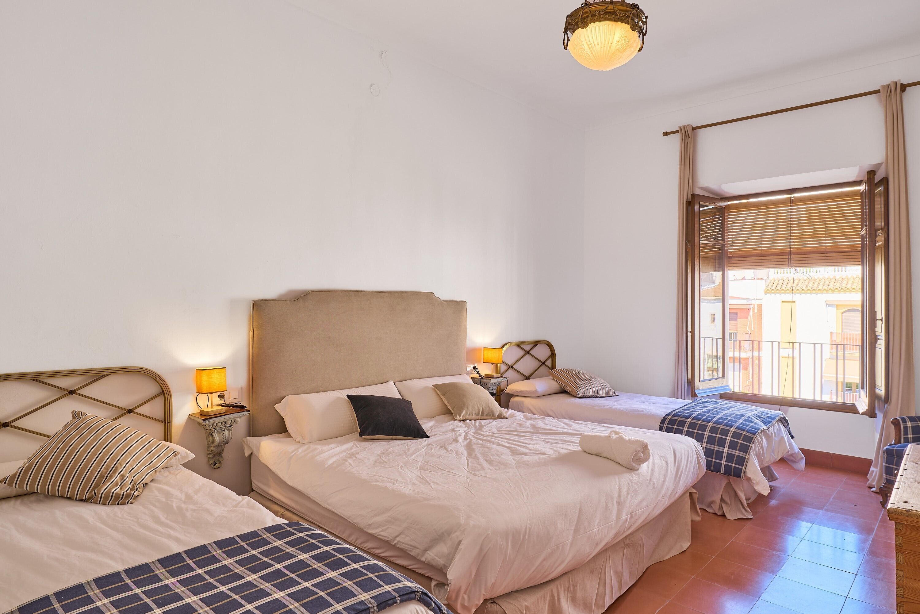 A hotel room with three beds, a chandelier, a window with blinds, and a view of the outside.