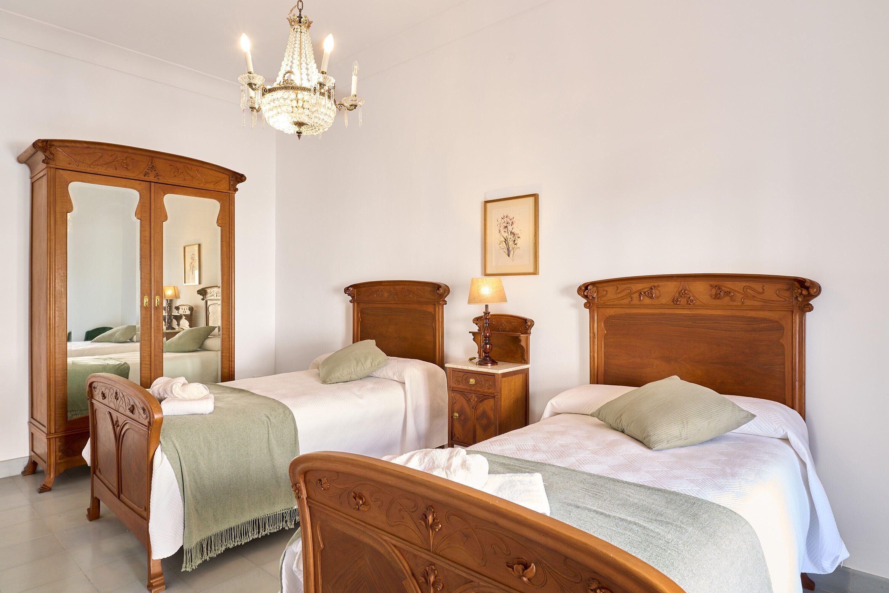 Un dormitorio con dos camas de madera, una mesita de noche con lámpara, un armario espejo y una araña.
