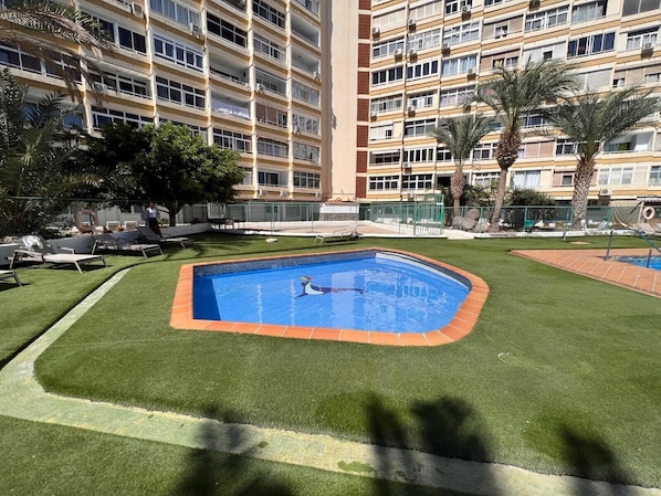 A swimming pool surrounded by artificial grass, with a multi-story building in the background.