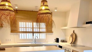 Een moderne keuken met een centraal eiland, houten krukken, hangende rieten kroonluchters en een raam met zonwering.