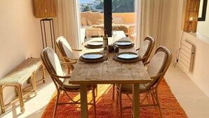 Een eetruimte met een houten tafel voor zes personen, omringd door rieten stoelen, een groot oranje vloerkleed, en uitzicht op de zee door een raam.