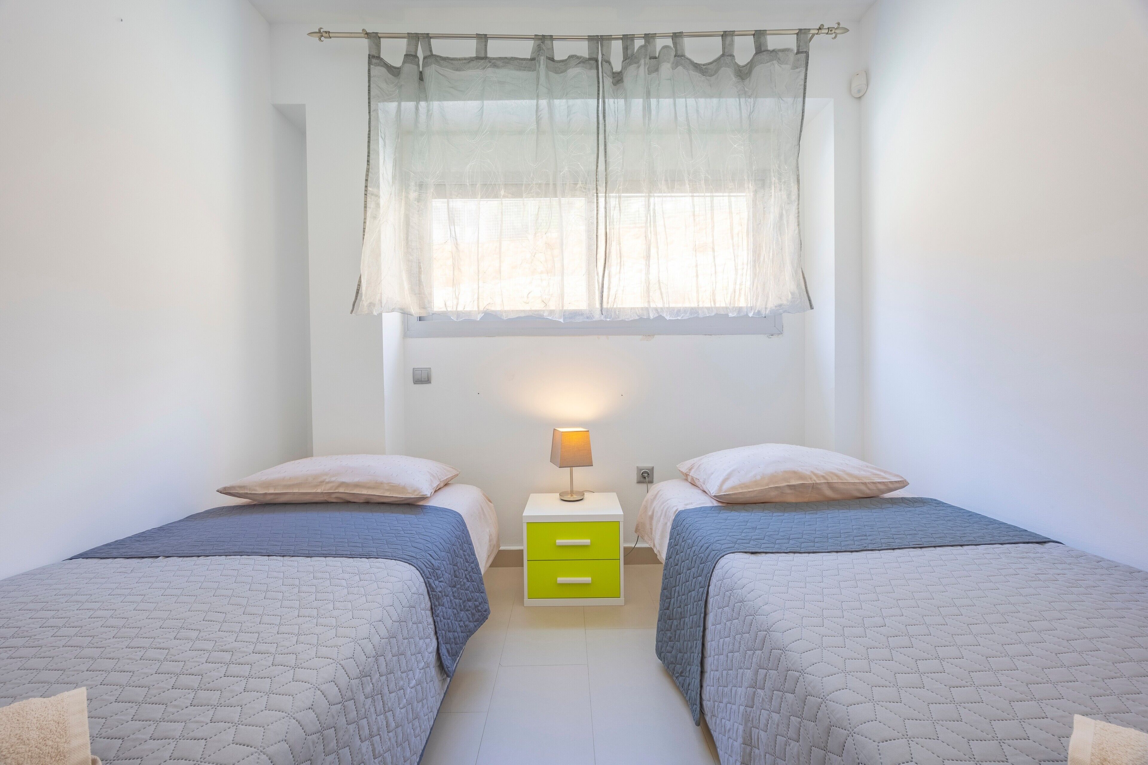 A room with two beds, a nightstand with a lamp, a window with sheer curtains, and a plain white wall.