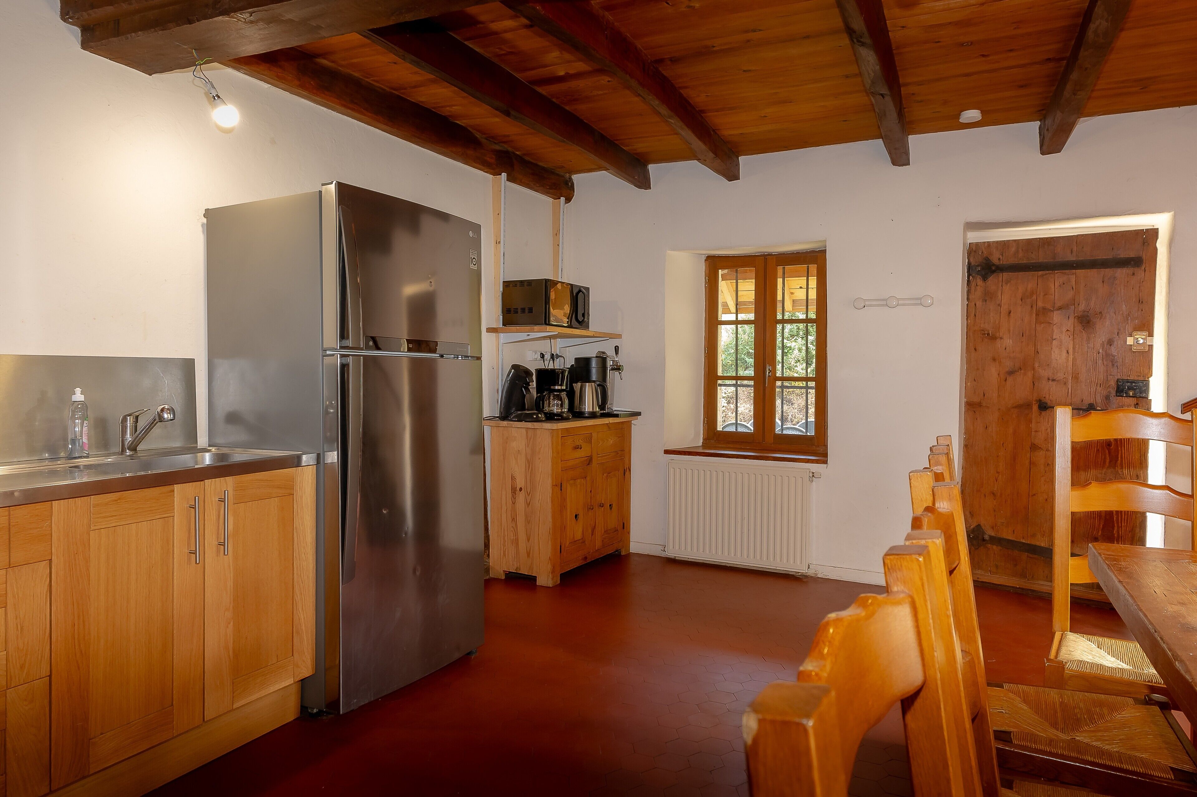 Una cocina con gabinetes de madera, un refrigerador de acero inoxidable, un mueble con electrodomésticos, una ventana y un comedor con sillas de madera.