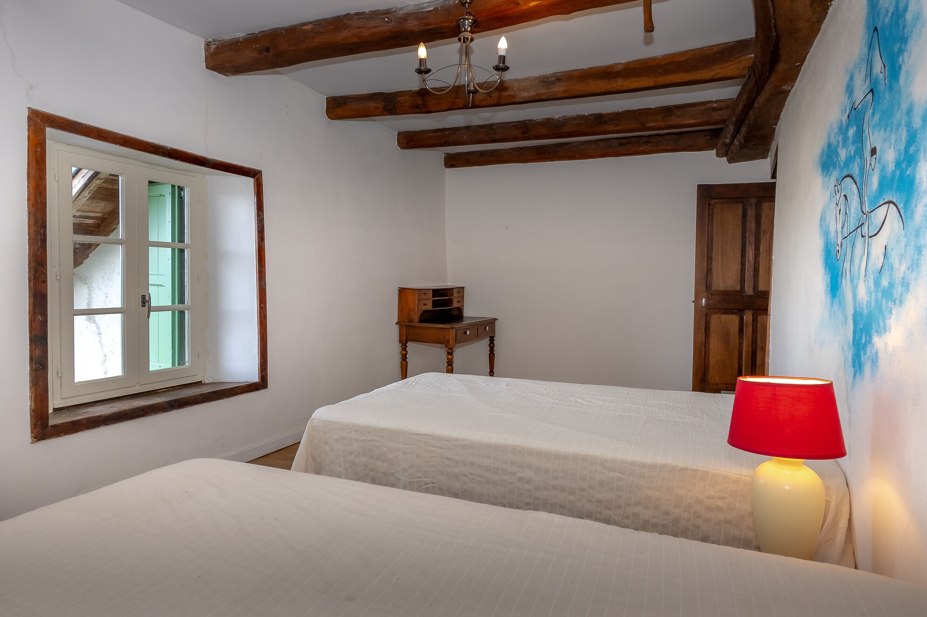 Une chambre avec deux lits, une chaise en bois, une lampe rouge, une fenêtre donnant sur un beau paysage et une poutre en bois au plafond.