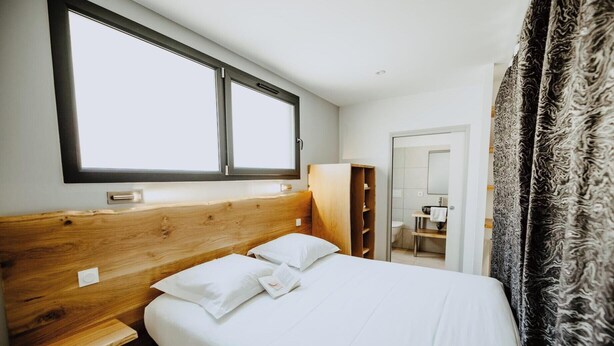 A bedroom with a wooden headboard, a large window, a wardrobe, and a bathroom visible through an open door. - Holiday Home 'Gite Villa T1' with Shared Pool, Wi-Fi and Air Conditioning (Vals-les-Bains)