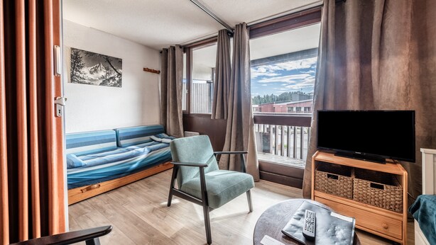Een kamer met een bed, een stoel, een tv-meubel, een raam met gordijnen en een schilderij aan de muur.