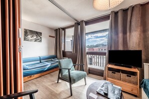Une pièce comprenant un lit, une chaise, un meuble pour la télévision, une fenêtre avec des rideaux et un tableau accroché au mur.