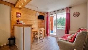 A cozy room with a wooden wall, a dining table with chairs, a sofa, and a window with curtains. - Apartment 'Appartement Alpe D'huez' with Balcony (Huez)