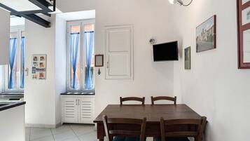 A dining area with a wooden table and chairs, a window with curtains, a television mounted on the wall, and a small window with a view of the outside.