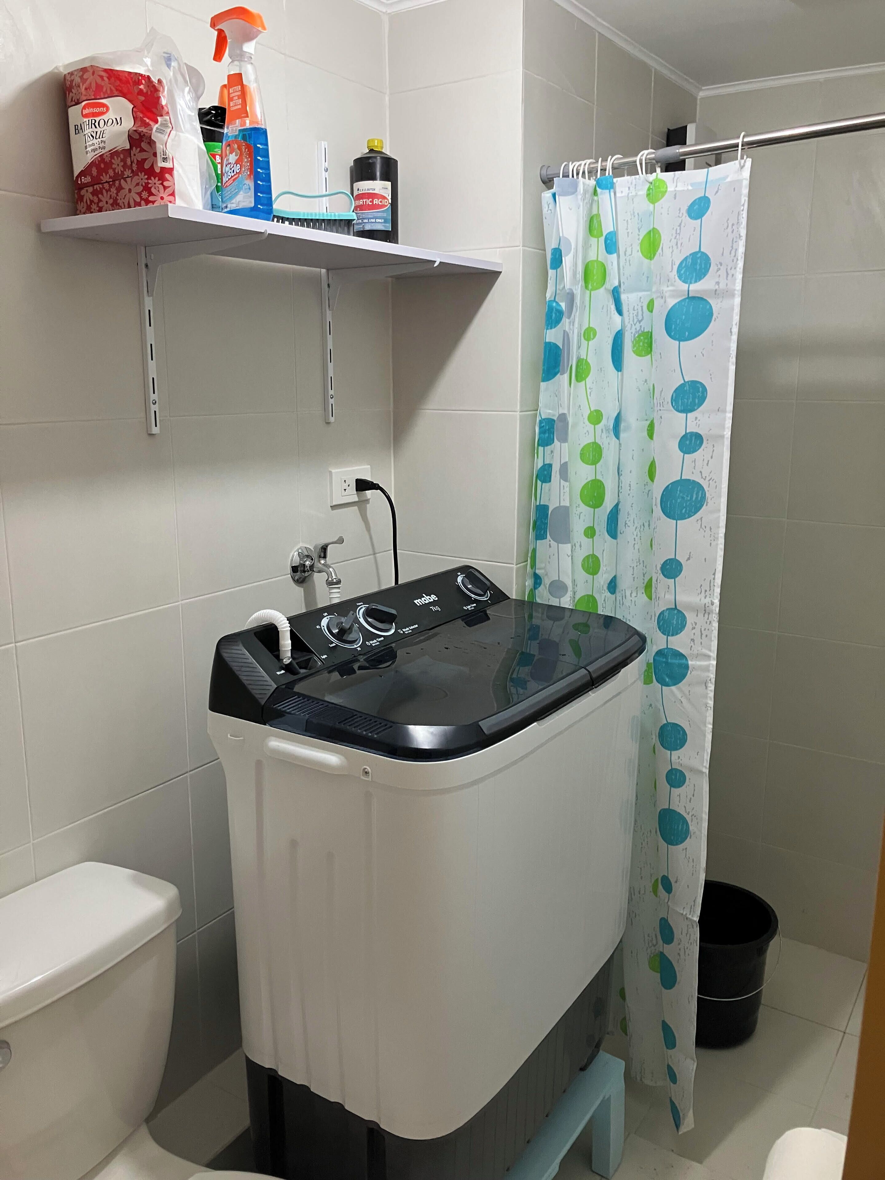 Un baño pequeño con lavadora, inodoro, un estante con productos de limpieza y cortina de ducha.