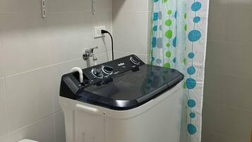 Un baño pequeño con lavadora, inodoro, un estante con productos de limpieza y cortina de ducha.