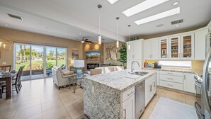 Una cocina amplia con una isla grande, muebles blancos y vista a un jardín ajardinado.