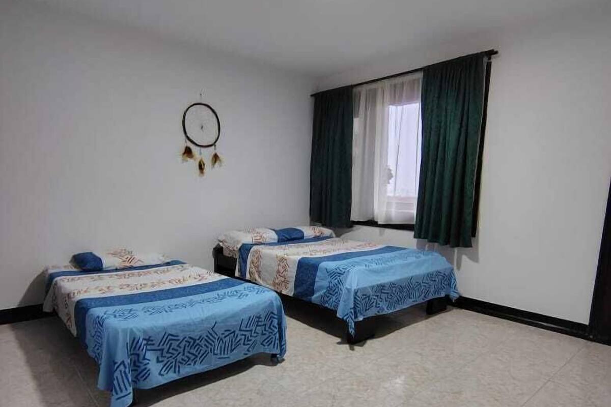 Una habitación con dos camas, un atrapasueños en la pared y una ventana con cortinas.