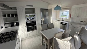 A compact kitchen with built-in appliances, a dining area with a table and chairs, and a window providing natural light. - Lakeside Caravan Sleeps 8 - Wifi, Parking, Views (Mablethorpe)