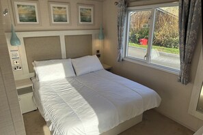 A neatly made bed in a room with a large window, a wall-mounted light fixture, and a power outlet. - Lakeside Caravan Sleeps 8 - Wifi, Parking, Views (Mablethorpe)