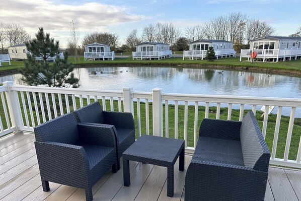 A deck with wicker furniture overlooking a lake, surrounded by mobile homes. - Lakeside Caravan Sleeps 8 - Wifi, Parking, Views (Mablethorpe)