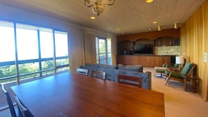 A spacious living area with a large wooden table and seating, a kitchen with modern appliances, and large windows offering a view of the outdoors. - A perfect view of Gabo Island. (Mallacoota)