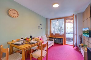 A dining area with a wooden table set for two, a clock on the wall, and a window with curtains.