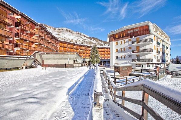 A snow-covered outdoor area with a wooden fence and a building with multiple balconies. - Studio Valleverde 4D, Sestriere, Italy (Sestriere)