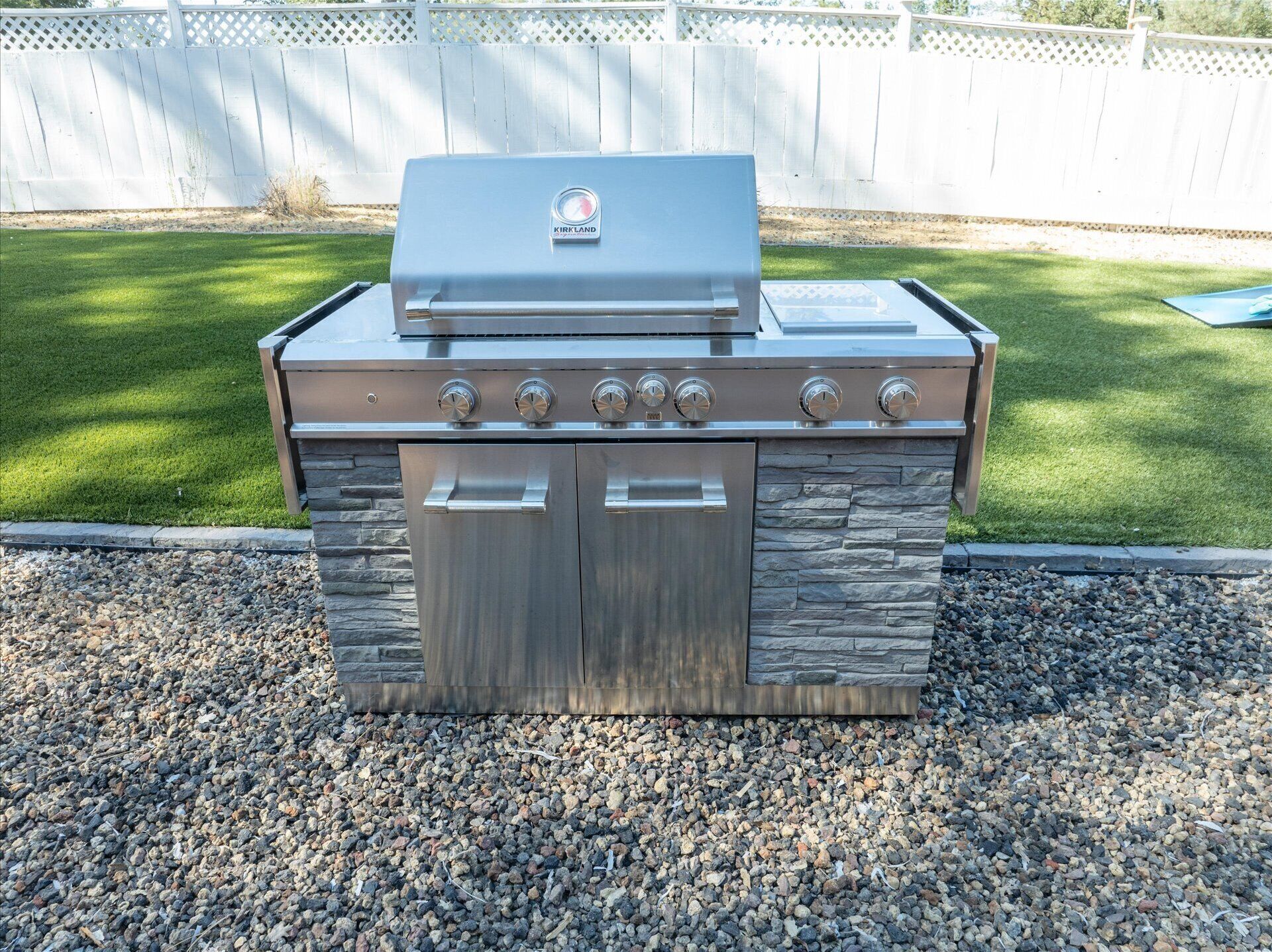 A stainless steel outdoor grill with a stone facade, located on a gravel surface with a grassy yard and a white fence in the background.