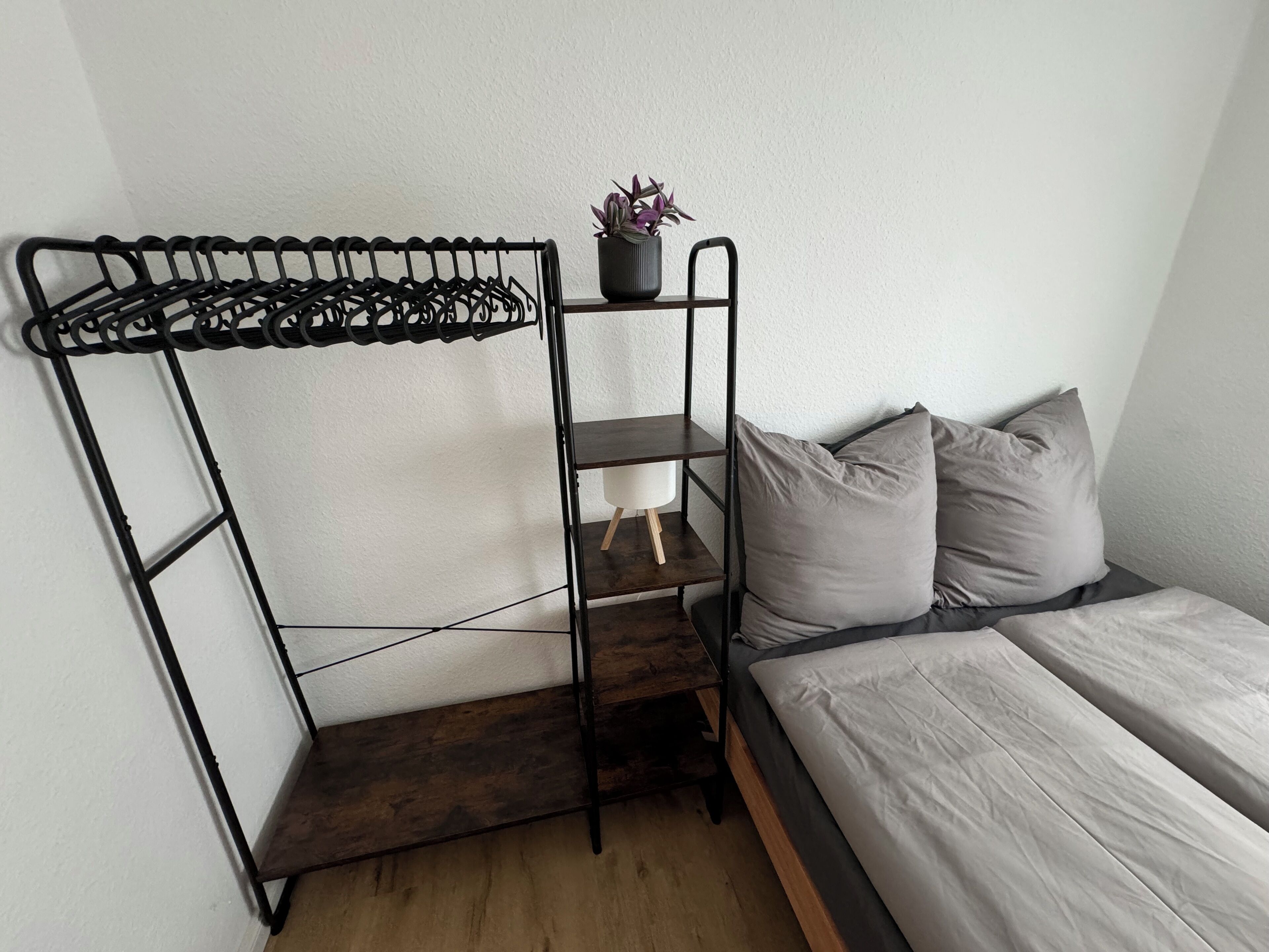 Una cama litera con estructura metálica, un estante pequeño con una planta en maceta, y una mesita de noche con lámpara.