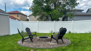 A backyard with two Adirondack chairs, a fire pit, and a white fence.