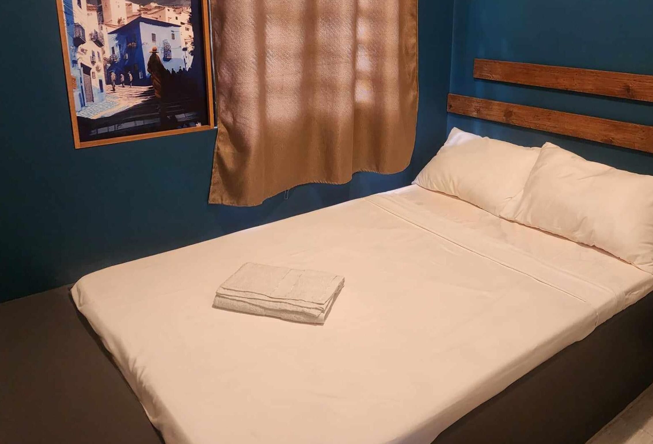 Una cama individual con colchón y almohadas color beige, cabecera de madera y un cuadro enmarcado con una vista urbana en la pared.