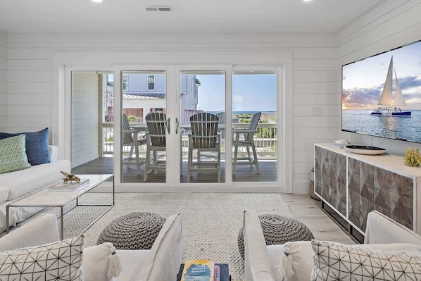 A modern living room with a sofa, coffee table, and TV, leading to a balcony with chairs and a sailboat view.