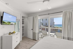 A bedroom with a TV, a bed, a balcony with a view of the sea, and a white ceiling fan.