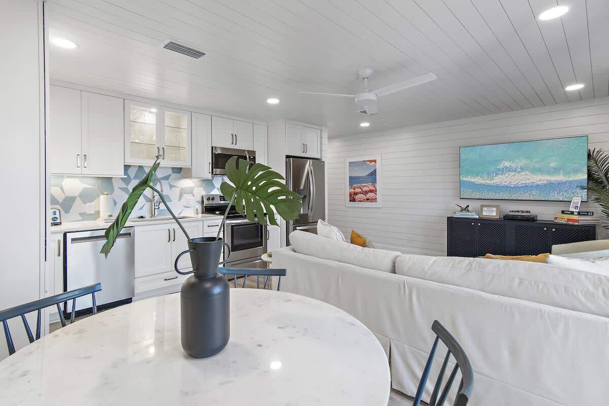 A modern kitchen with white cabinets, a dining table with chairs, a large plant, and a television on a black stand.