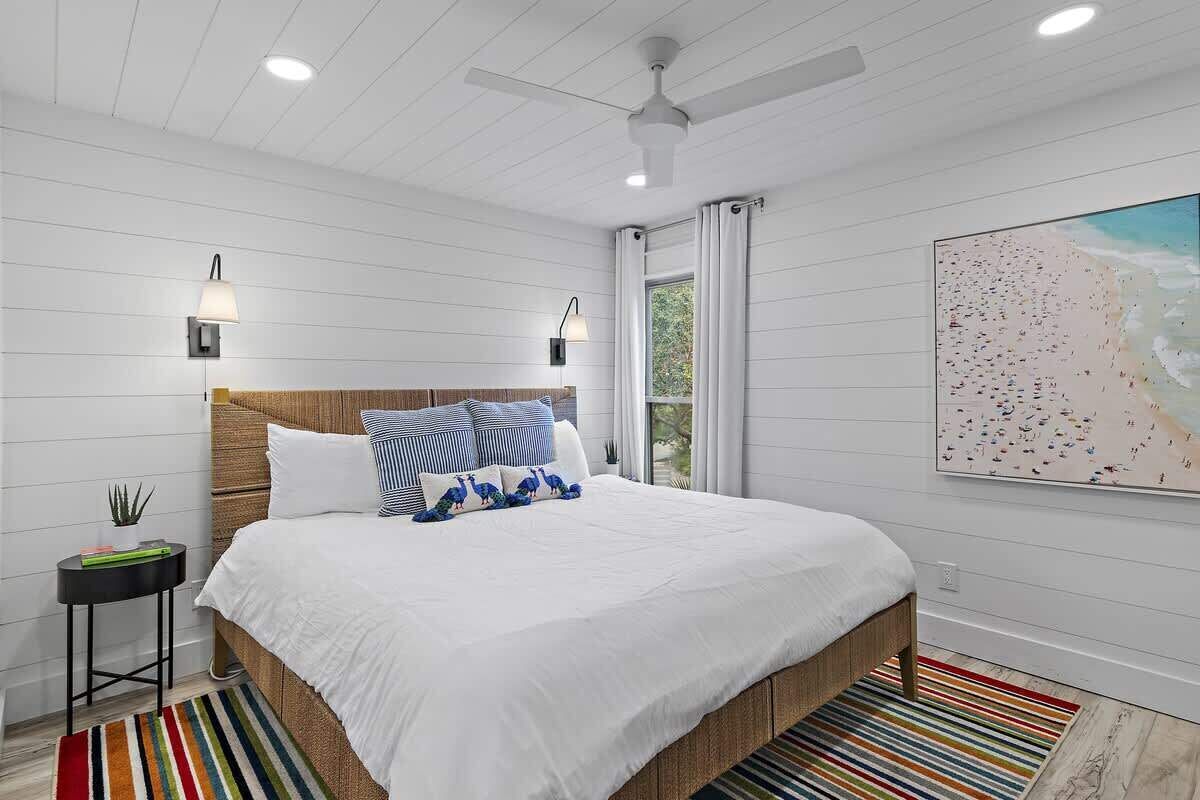 A bedroom with a bed, a ceiling fan, two wall-mounted lamps, a window with curtains, and a beach-themed painting.