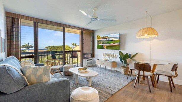Ein geräumiges Wohnzimmer mit einer großen Schiebeglastür, die auf einen Balkon führt, einem runden Esstisch mit Stühlen, einem Sofa mit Kissen, einem Couchtisch und einem Deckenventilator.