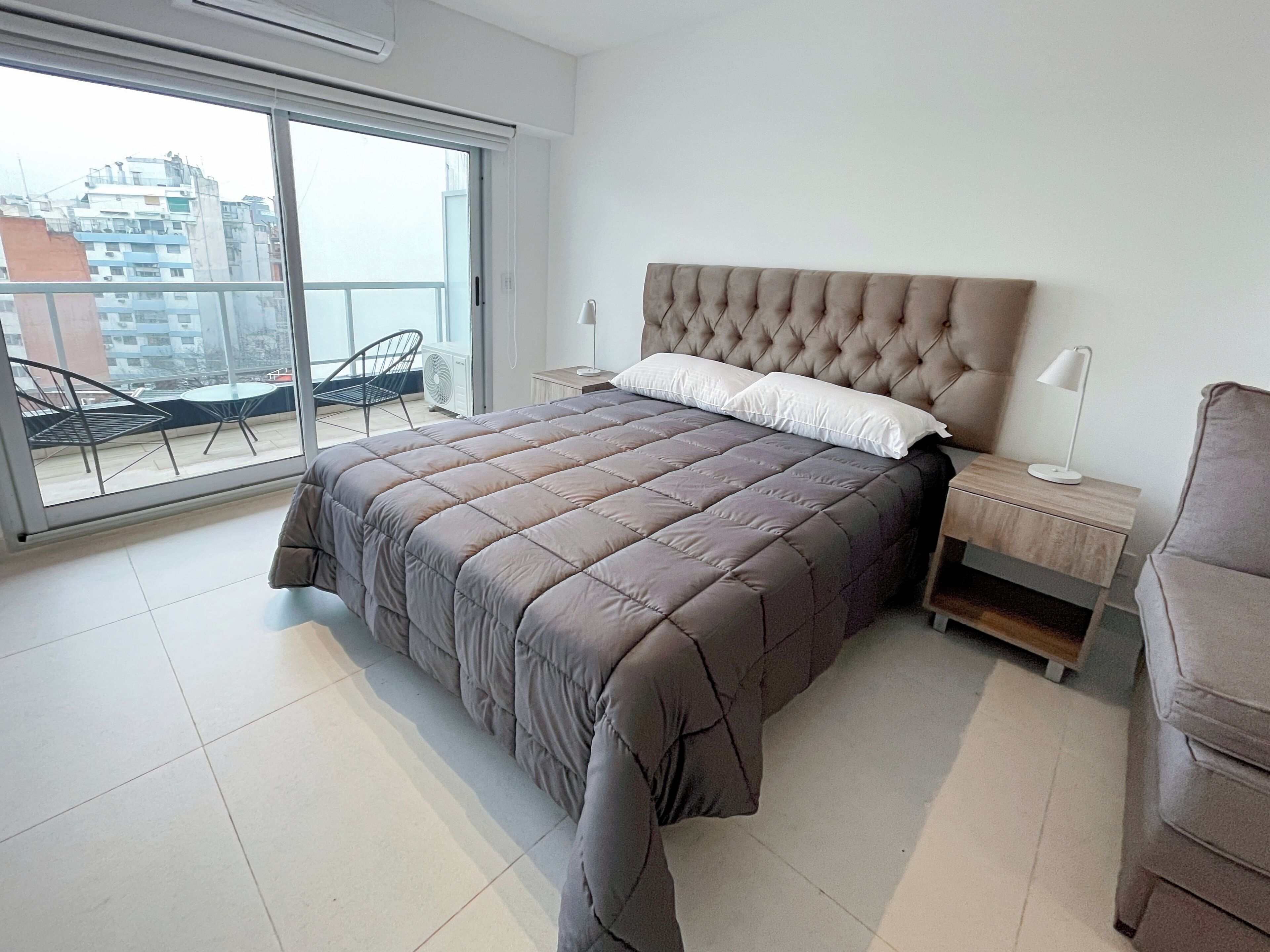 A bedroom with a large bed, a nightstand, a chair, and a view of the city through a sliding door.