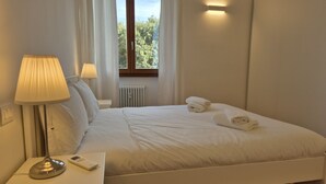 Ein ordentlich bezogenes Bett mit weißen Bettwäsche und Handtüchern, eine Nachttischlampe und Blick auf Bäume durch ein Fenster.