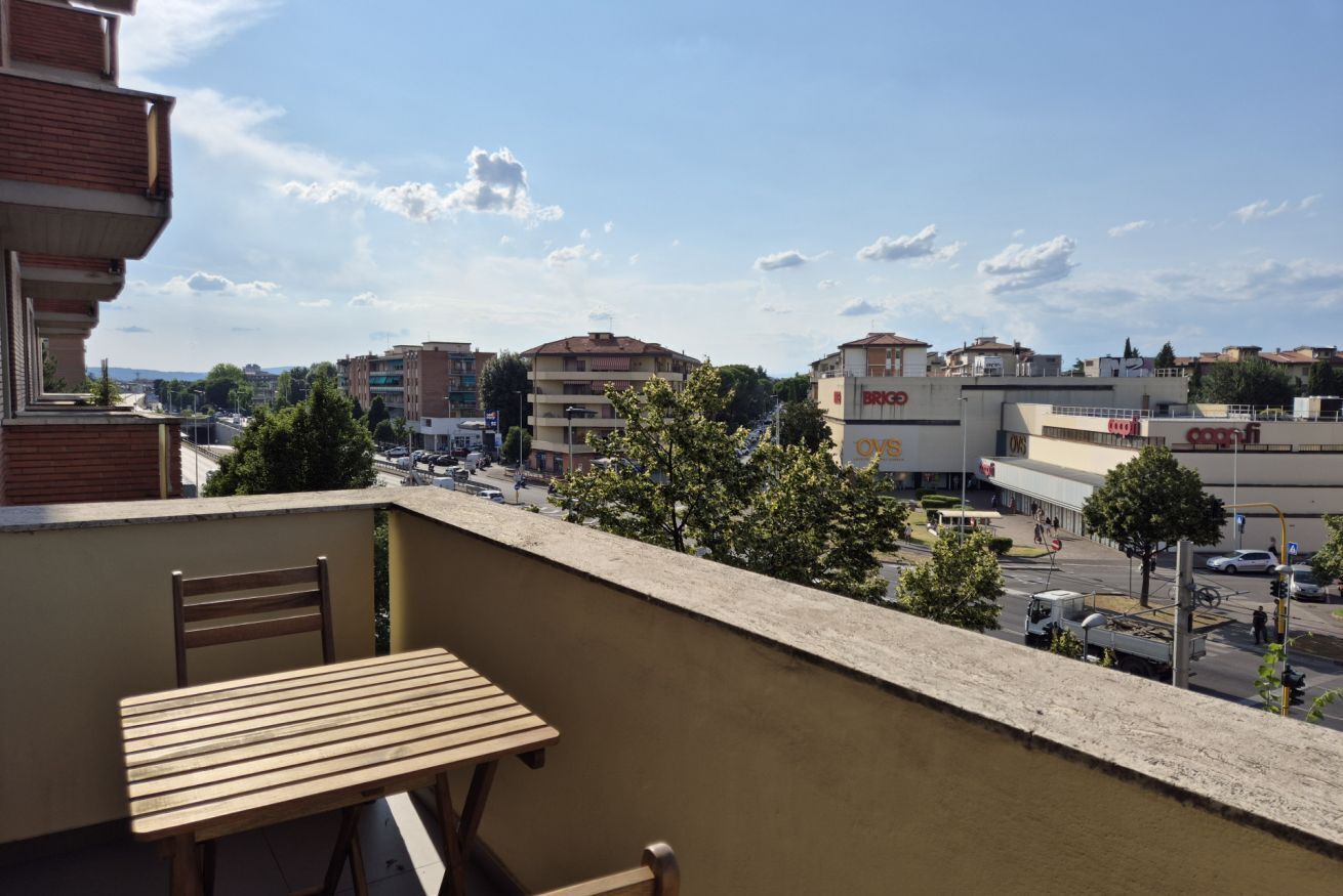 A balcony with a wooden table and chairs, overlooking a cityscape with various buildings and a clear sky.