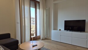 Ein modernes Wohnzimmer mit einer Couch, einem runden Couchtisch, einem Fernseher auf einem weißen Schrank und einer Glastür, die auf einen Balkon führt.