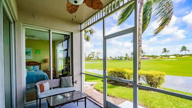 A room with a large sliding glass door leading to a golf course view, a bed with a blue headboard, and a small table with a bowl. - Stunning 3BR Naples Condo with Tropical Views (Naples)