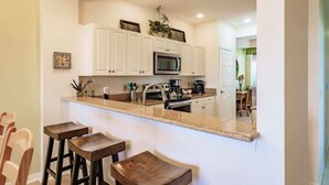 A kitchen with white cabinets, a microwave, a sink, and a countertop. There are three wooden stools and a plant on the countertop. - Stunning 3BR Naples Condo with Tropical Views (Naples)