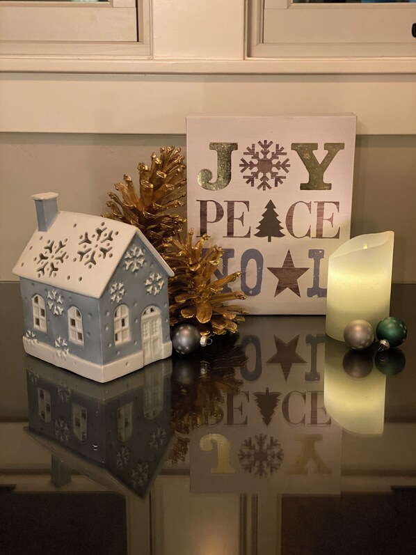 A small blue house-shaped decoration with snowflakes, a sign that reads "JOY PEACE TO ALL," and a candle on a reflective surface.