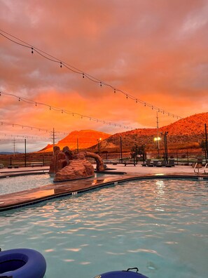 A pool area with a rock formation, lounge chairs, and string lights against a sunset backdrop.